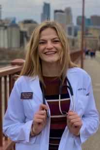 Olivia Wicker wearing her whitecoat, smiling toward the camera while standing on the Stone Arch Bridge in Minneapolis