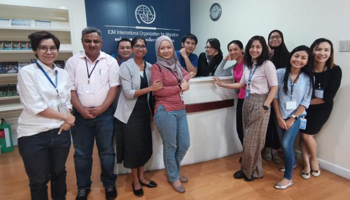 A group of mostly women standing and smiling in front of a desk with IOM International Organization of Migration visible on the wall in the background.